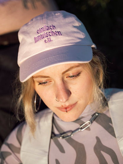 Person mit Piercings trägt helle Cap mit lila „einfach knutschen e.V.“ Stickerei im Abendlicht.